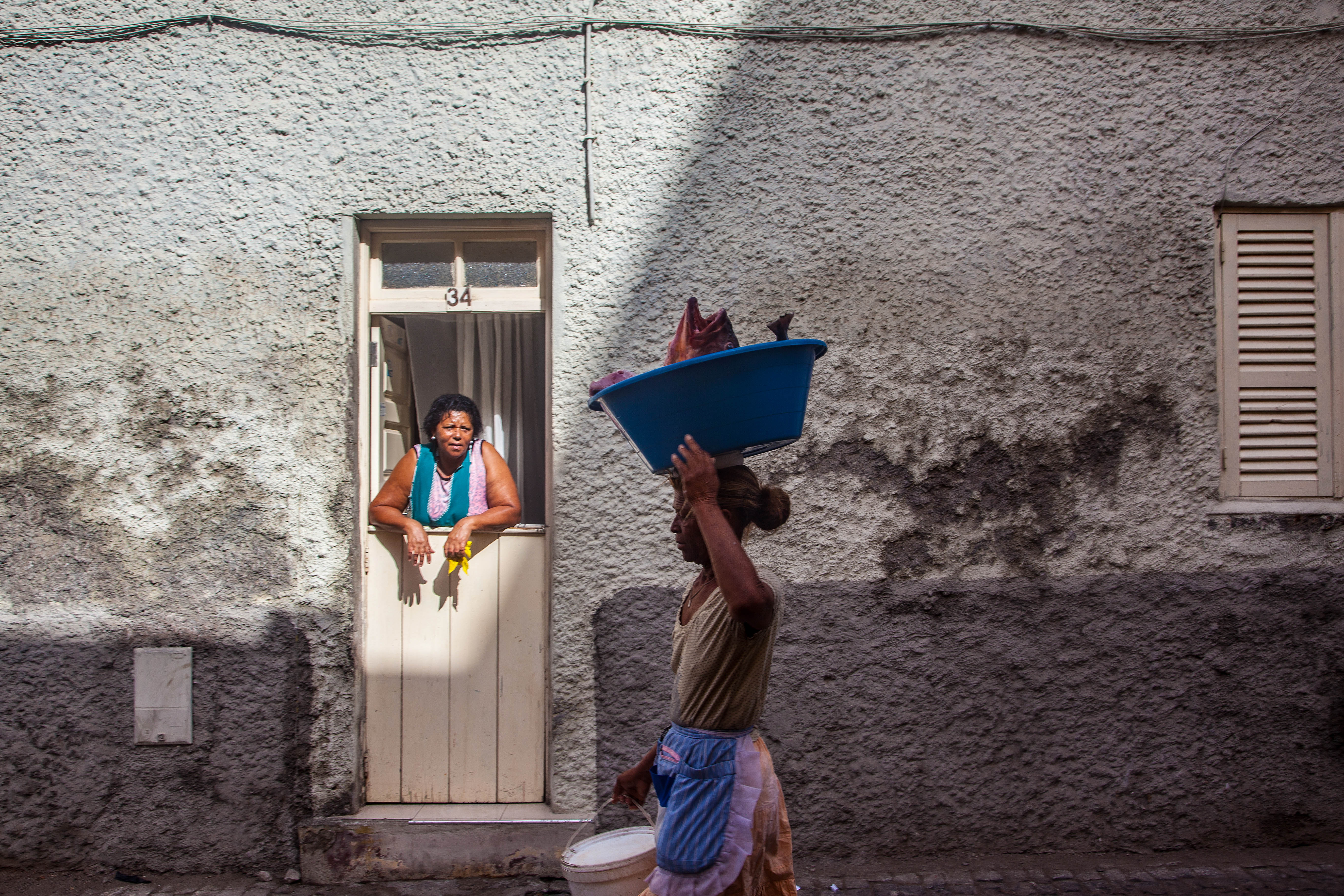 Vrouw loopt door straat met kom met grote viskop in op het hoofd terwijl een dame uit haar deur leunend haar nakijkt